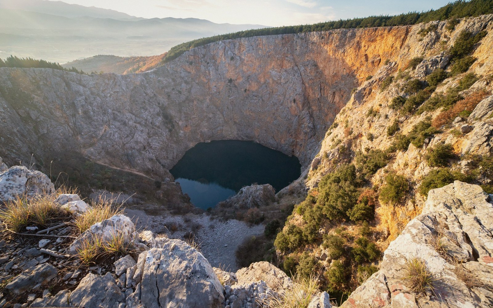 Steep limestone sinkhole with a dark lake at its base surrounded by forested cliffs